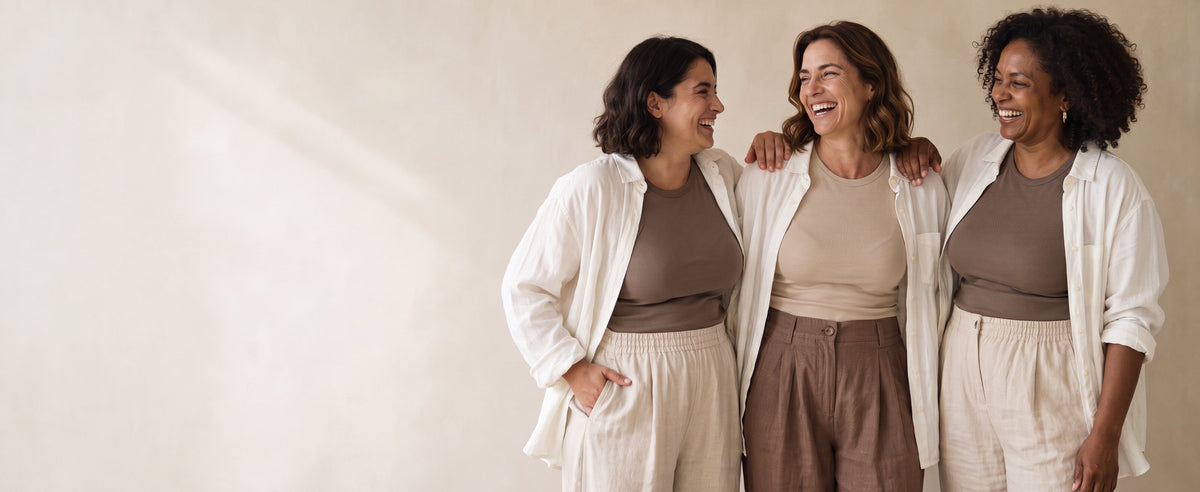 Trois femmes aux silhouettes généreuses et variées, en chemises de lin claires et pantalons souples, souriant ensemble sur fond beige